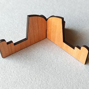 May include: A wooden phone stand with a natural wood finish and dark brown edges. The stand is designed with a unique shape, featuring a central hinge and two angled support arms. The stand is designed to hold a phone or small tablet.