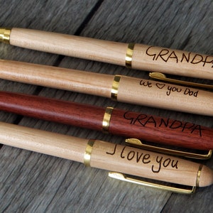 May include: Four ballpoint pens with wooden barrels and gold-tone metal accents. Three pens are light-colored wood, one is dark reddish-brown wood. Engraved text includes "GRANDPA", "we heart you Dad", "GRANDPA", and "I love you" on the barrels.