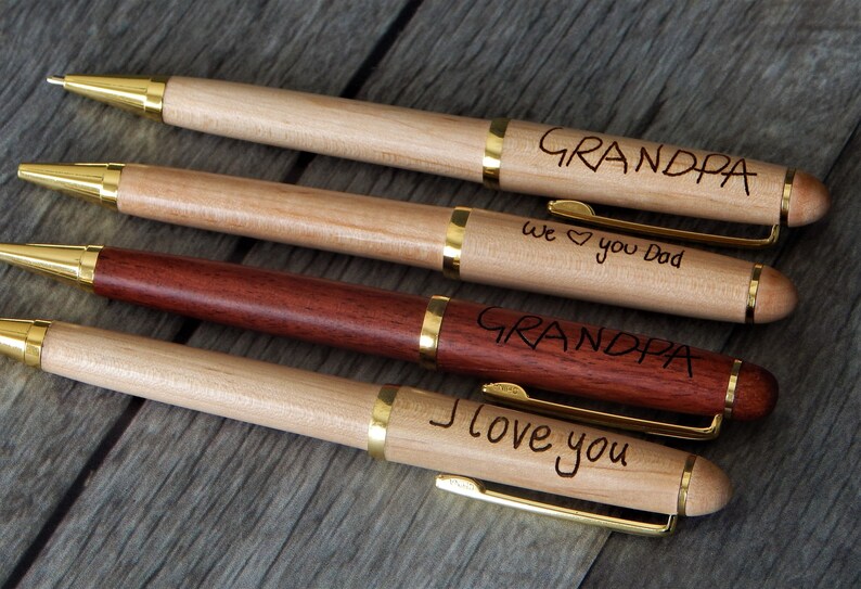 May include: Four ballpoint pens with wooden barrels and gold-tone metal accents. Three pens are light-colored wood, one is dark reddish-brown wood. Engraved text includes "GRANDPA", "we heart you Dad", "GRANDPA", and "I love you" on the barrels.