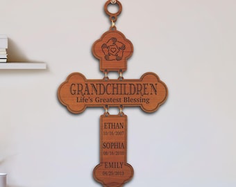 May include: A brown wooden wall hanging shaped like a cross, featuring a top section with a hand-holding-heart icon, a center bar engraved with "GRANDCHILDREN Life's Greatest Blessing," and a vertical piece listing names Ethan, Sophia, and Emily.