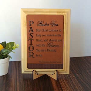 May include: A framed plaque with a brown leather-like insert. The text reads "Pastor Van" and a religious message. The frame is light brown, and the plaque is displayed on a wooden stand. The text is in black.