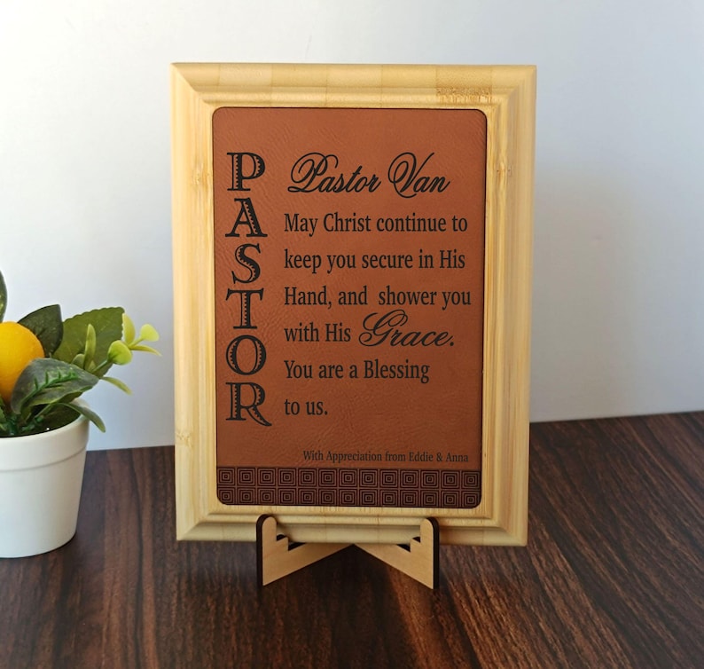 May include: A framed plaque with a brown leather-like insert. The text reads "Pastor Van" and a religious message. The frame is light brown, and the plaque is displayed on a wooden stand. The text is in black.