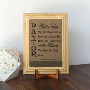 May include: A framed plaque with a light brown wooden frame and a brown leather-like insert. The text reads "Pastor Van" and a message of appreciation. The plaque is displayed on a wooden stand.