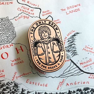 May include: A copper-colored pin with a cartoon illustration of a person wearing a brown jacket and a hat. The pin is oval-shaped and has the text "The Only Brew For The Brave & True" around the edge.