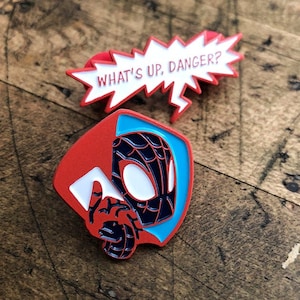 May include: Two enamel pins featuring Spider-Man designs. One pin is a red and white speech bubble with the text "WHAT'S UP, DANGER?". The other pin depicts Spider-Man's mask and hand, with red, blue, and black colors.
