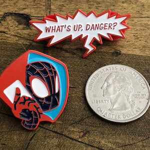 May include: Enamel pins featuring Spider-Man designs. One pin shows Spider-Man's face and hand, another is a speech bubble with "WHAT'S UP, DANGER?" in white text. A quarter is included for size reference.