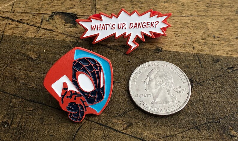 May include: Enamel pins featuring Spider-Man designs. One pin shows Spider-Man's face and hand, another is a speech bubble with "WHAT'S UP, DANGER?" in white text. A quarter is included for size reference.