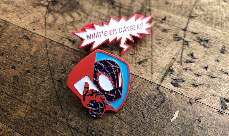 May include: Two enamel pins featuring Spider-Man designs. One pin is a red and white speech bubble with the text "WHAT'S UP, DANGER?". The other pin depicts Spider-Man's mask and hand, with red, blue, and black colors.