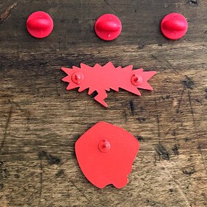 May include: A collection of red enamel pins on a wooden surface. The pins include three circular pins, a speech bubble pin, and a square pin with a rounded edge. The pins are arranged on a textured, brown wooden surface.