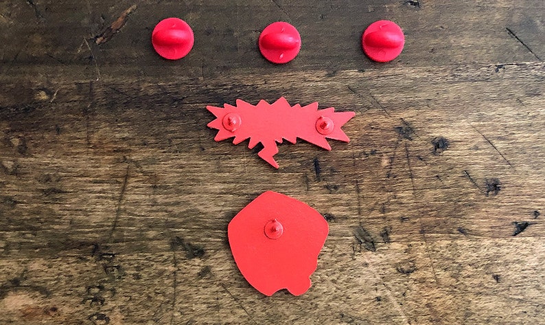 May include: A collection of red enamel pins on a wooden surface. The pins include three circular pins, a speech bubble pin, and a square pin with a rounded edge. The pins are arranged on a textured, brown wooden surface.