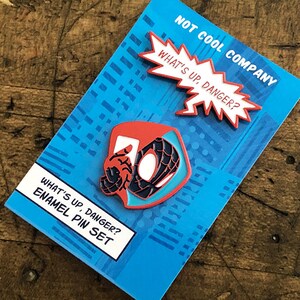May include: Enamel pin set featuring a red, black, and blue Spider-Man design with the text "WHAT'S UP, DANGER?" and "NOT COOL COMPANY" on a blue background. The set includes two pins.