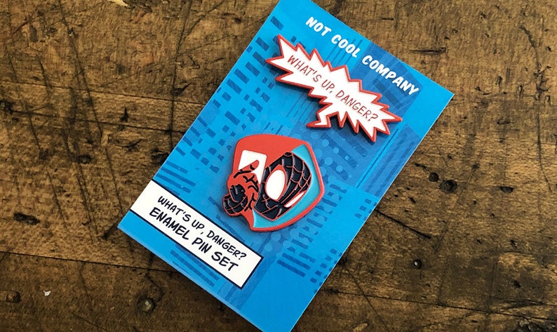 May include: Enamel pin set featuring a red, black, and blue Spider-Man design with the text "WHAT'S UP, DANGER?" and "NOT COOL COMPANY" on a blue background. The set includes two pins.