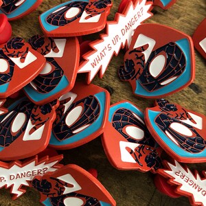 May include: A collection of red, blue, and white enamel pins. The pins feature a stylized Spider-Man mask design and the text "WHAT'S UP, DANGER?" in a speech bubble. The pins are scattered on a wooden surface.
