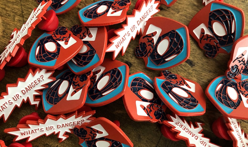 May include: A collection of red, blue, and white enamel pins. The pins feature a stylized Spider-Man mask design and the text "WHAT'S UP, DANGER?" in a speech bubble. The pins are scattered on a wooden surface.