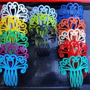 May include: A collection of colorful plastic hair combs with a decorative, ornate design. The combs are arranged in a stack and are all different colors, including white, yellow, green, black, orange, red, and blue.