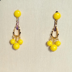 May include: A pair of yellow dangle earrings. Each earring features a large yellow bead at the top, a gold-colored decorative element with a purple accent, and three smaller yellow beads hanging below. The earrings are displayed against a light background.