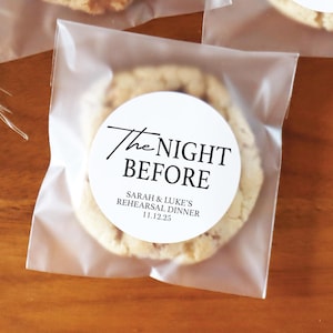 May include: Three cookies in clear cellophane bags, each with a white circular label. The labels read "The Night Before" and "Sarah & Luke's Rehearsal Dinner 11.12.25" in black script. The cookies are on a wooden surface.