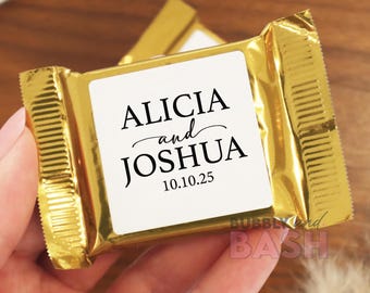 May include: A gold foil-wrapped chocolate bar with a white label featuring the names "ALICIA and JOSHUA" and the date "10.10.25" in black script. The bar is held in a hand, suggesting a wedding favor or celebratory treat.