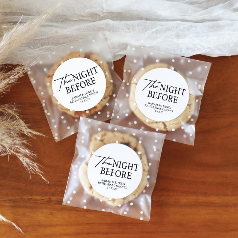 May include: Three cookies in clear cellophane bags with white polka dots. Each bag has a circular sticker that reads "The Night Before" with event details. The cookies are on a wooden surface, suggesting a wedding or rehearsal dinner.