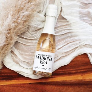 May include: A small bottle of sparkling wine with a white label that says "Welcome to your Madrina Era Are you ready for it?" with a heart symbol.