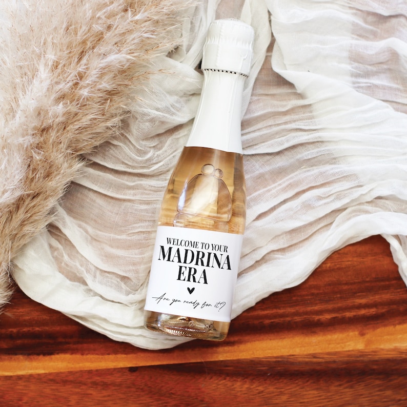 May include: A small bottle of sparkling wine with a white label that says "Welcome to your Madrina Era Are you ready for it?" with a heart symbol.