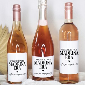 May include: Three wine bottles with labels that say "Welcome to your Madrina Era. Are you ready for it?" The labels are white with black text and a small heart.