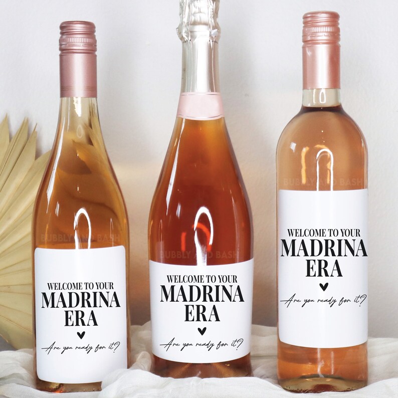 May include: Three wine bottles with labels that say "Welcome to your Madrina Era. Are you ready for it?" The labels are white with black text and a small heart.