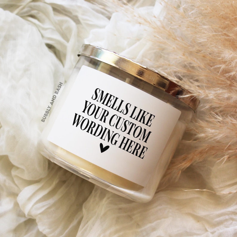 May include: A clear glass candle jar with a silver lid and a white label that reads "Smells Like Your Custom Wording Here" with a small black heart.