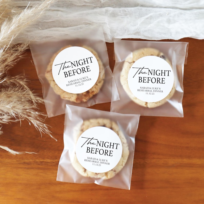 May include: Three cookies in clear cellophane bags, each with a white circular label. The labels read "The Night Before" and "Sarah & Luke's Rehearsal Dinner 11.12.25" in black script. The cookies are on a wooden surface.