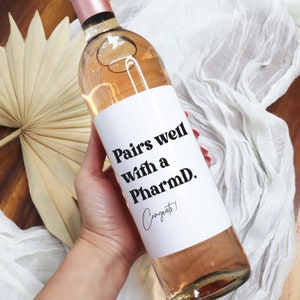 May include: A white wine bottle label with black text that reads "Pairs well with a PharmD. Congrats!"