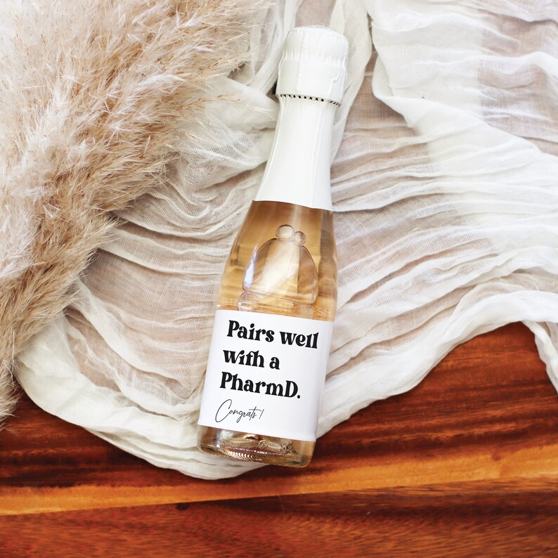 May include: A small bottle of champagne with a white label that says "Pairs well with a PharmD. Congrats!" The bottle is sitting on a wooden surface with a white fabric background.