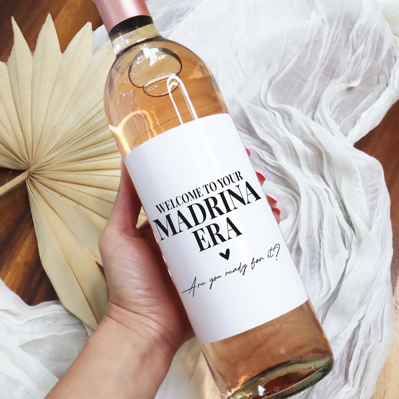 May include: A white wine bottle with a black and white label that says "Welcome to your Madrina Era" with a black heart and the text "Are you ready for it?" in cursive.