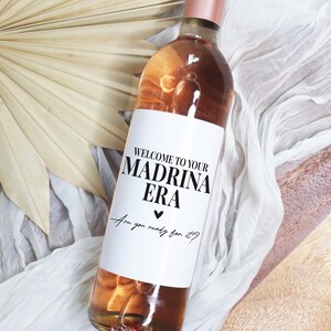 May include: A white wine bottle with a label that says "Welcome to your Madrina Era. Are you ready for it?" The label has a black heart.