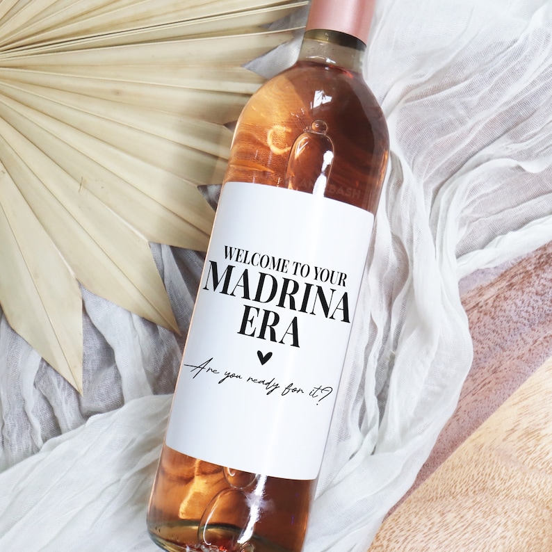 May include: A white wine bottle with a label that says "Welcome to your Madrina Era. Are you ready for it?" The label has a black heart.