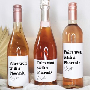 May include: Three bottles of wine with white labels that say "Pairs well with a PharmD. Congrats!" The bottles are a light pink color.