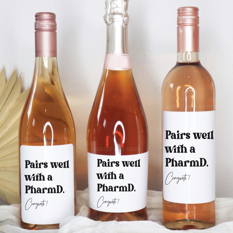 May include: Three bottles of wine with white labels that say "Pairs well with a PharmD. Congrats!" The bottles are a light pink color.