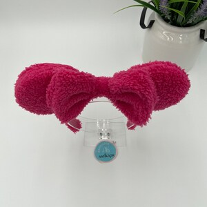 May include: A pink fluffy headband with a large bow. The headband is on a white background with a small blue tag that says "Handmade by Antikapr".