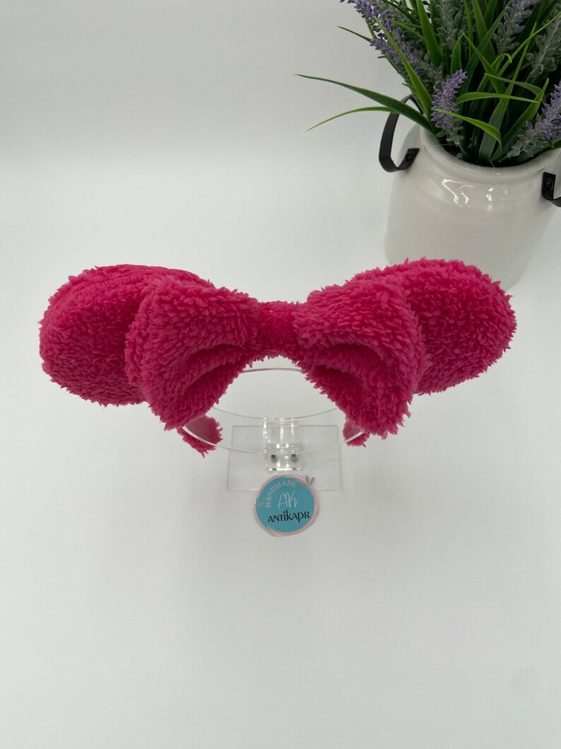 May include: A pink fluffy headband with a large bow. The headband is on a white background with a small blue tag that says "Handmade by Antikapr".