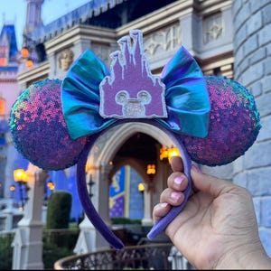 Puede incluir: Orejas de Minnie Mouse iridiscentes de color morado y azul con un lazo en forma de castillo morado. El castillo tiene un contorno blanco y una silueta blanca de la cabeza de Mickey Mouse en su interior.