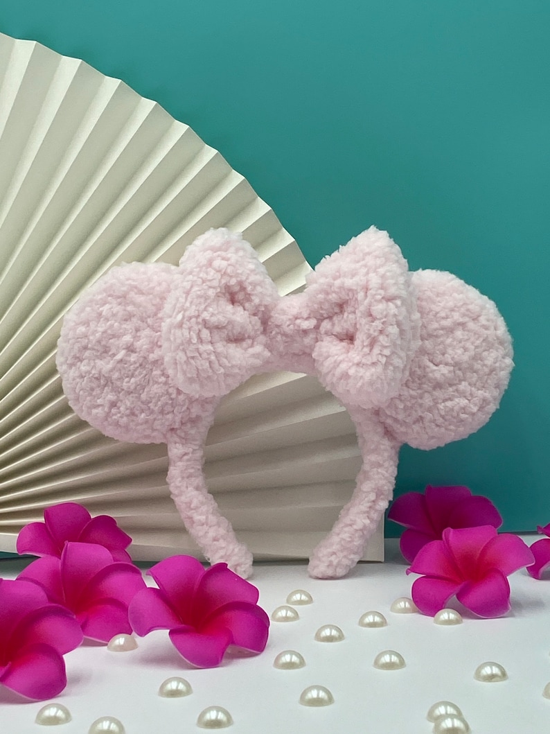 Pu&ograve; includere: Un cerchietto rosa peloso con grandi orecchie soffici e un fiocco al centro. Il cerchietto &egrave; su una superficie bianca con fiori rosa e perle bianche.