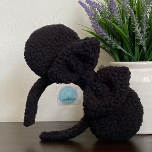 May include: Black fuzzy headband with large, round, fuzzy ears. The headband is shaped like Mickey Mouse ears. The headband has a small, white tag with the text "Handmade by Anikapr" on it.