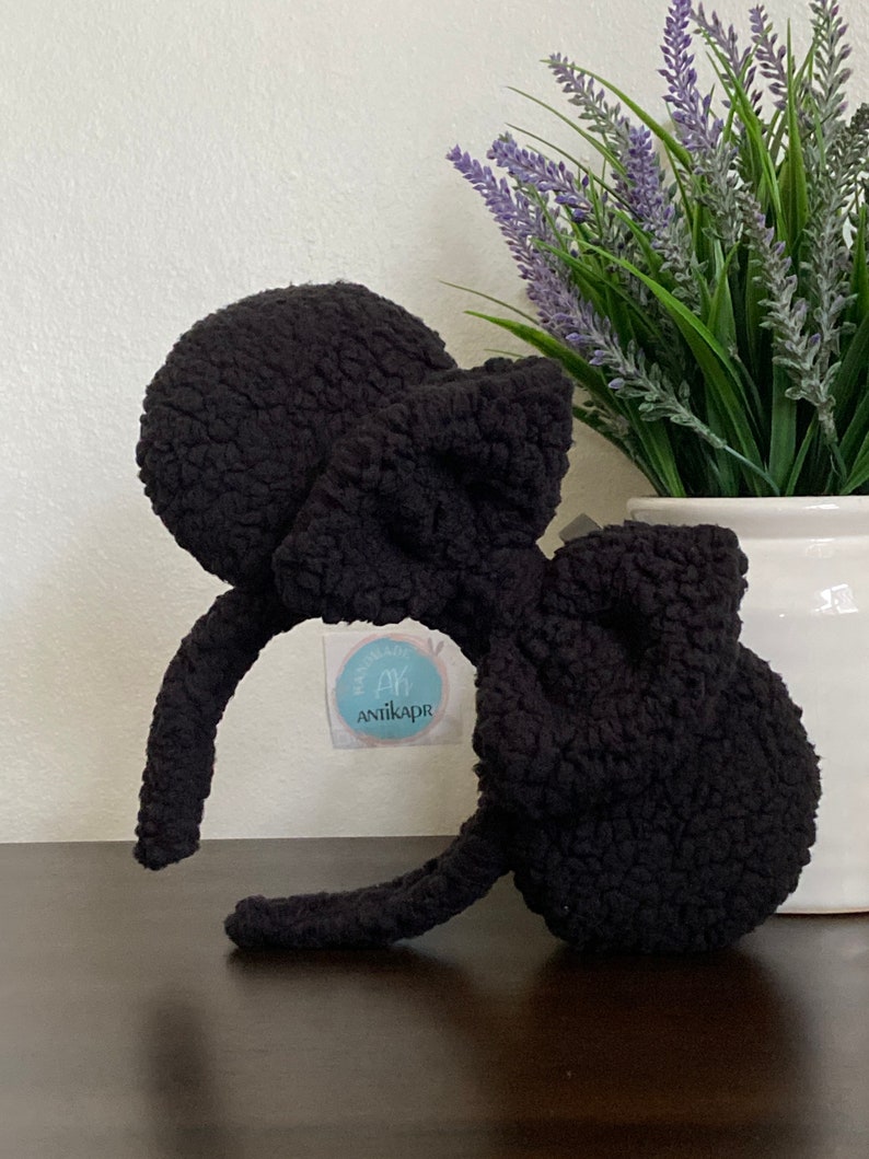 May include: Black fuzzy headband with large, round, fuzzy ears. The headband is shaped like Mickey Mouse ears. The headband has a small, white tag with the text "Handmade by Anikapr" on it.