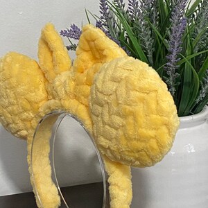 May include: Yellow plush Minnie Mouse ears headband with a woven texture. The headband is on a clear stand.