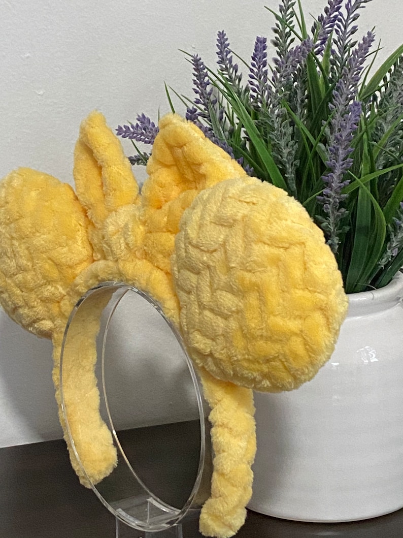 May include: Yellow plush Minnie Mouse ears headband with a woven texture. The headband is on a clear stand.