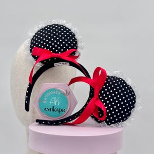 May include: A pair of black headbands with round ears featuring a white polka dot pattern, white lace trim, and a bright red satin bow on each. A hexagonal light purple sign with the text HANDMADE AK ANTIKAPR is positioned between the headbands.
