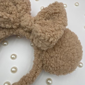 May include: A brown fuzzy headband with large bow and ear-like accents. The headband is made of a soft, plush material.