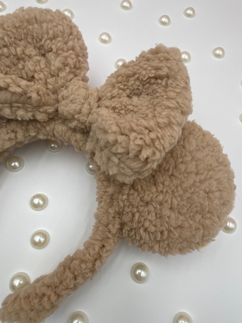 May include: A brown fuzzy headband with large bow and ear-like accents. The headband is made of a soft, plush material.