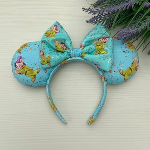 May include: Light blue Minnie Mouse ears headband with a large bow and a print of the Seven Dwarfs from Snow White. The ears are covered in a light blue glitter fabric with a white confetti pattern.