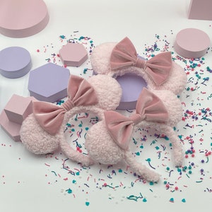 May include: Two pink fuzzy mouse ear headbands with pink velvet bows. The headbands are on a white surface with pink, blue and green sprinkles.