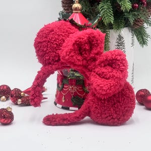 Puede incluir: Orejeras rojas y difusas con una diadema. Las orejeras est&aacute;n hechas de un material suave y afelpado. Adornos navide&ntilde;os rojos y un globo de nieve decoran el fondo.